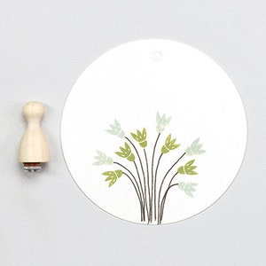 Könnte beinhalten: Ein runder weißer Anhänger mit einem Loch oben und einem grünen Blumenmuster. Das Muster zeigt eine Gruppe von stilisierten grünen und blauen Blumen mit schwarzen Stielen.