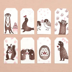 May include: Set of eight white gift tags with brown animal illustrations. The tags feature a meerkat, seal, birds, raccoon, squirrel, moles, hedgehog, and dinosaur. Each tag has a unique design with pink accents, perfect for gifts.