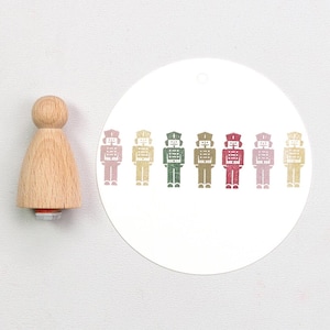 Könnte beinhalten: Ein Holzstempel mit Griff neben einem weißen kreisförmigen Anhänger. Der Anhänger zeigt ein sich wiederholendes Muster aus bunten Spielzeugsoldatenfiguren in Rosa, Gelb, Grün und Rot. Der Stempel ist aus hellem Holz gefertigt.