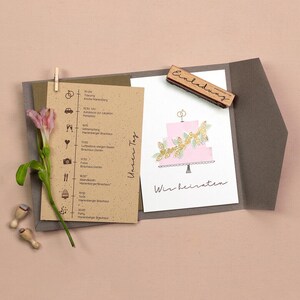 May include: A wedding invitation suite featuring a pink cake illustration with gold leaf accents. The invitation includes a schedule of events and the words "Wir heiraten". A wooden stamp with the word "Einladung" is also present.