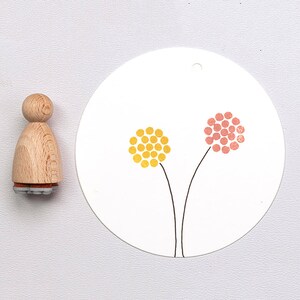 Op de afbeelding: Een houten stempel en een wit label met een bloemmotief. De stempel heeft een licht houten handvat en een rubberen basis met patroon. Het label toont twee gestileerde bloemhoofden in geel en roze.