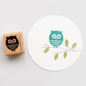 May include: A wooden stamp featuring a black owl design, alongside a white circular tag adorned with a teal owl illustration on a branch with green leaves. The stamp has the text "© PERLENFISCHER 2014".