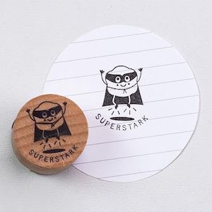 Könnte beinhalten: Ein Holzstempel mit einem Cartoon-Superhelden-Design und dem Wort "SUPERSTARK" aufgedruckt. Der Stempel befindet sich neben einem weißen Papierkreis mit dem gleichen Design. Der Superheld trägt eine Maske und einen Umhang.