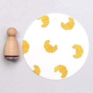 Könnte beinhalten: Ein runder, weißer Papiersticker mit einem sich wiederholenden Muster aus gelben Halbmondformen. Der Sticker hat einen Durchmesser von 7,6 cm. Im Hintergrund befindet sich ein Stempel aus Holz mit einer Personenform.