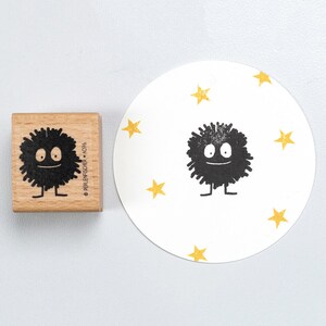 Könnte beinhalten: Ein Holzstempel mit einem Cartoon-Charakter-Design und einem gestempelten Bild auf einem weißen Kreis. Der Stempel ist ein quadratischer Block mit einem schwarzen, stacheligen Charakter mit einem lächelnden Gesicht und kleinen Beinen. Das gestempelte Bild ist von gelben Sternen umgeben.