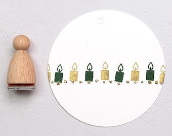 Pode incluir: Um carimbo de madeira com um desenho de vela ao lado de uma etiqueta branca com uma borda de vela. As velas alternam entre ouro e verde, com pequenos detalhes dourados por baixo. O carimbo tem um cabo de madeira e uma base vermelha.
