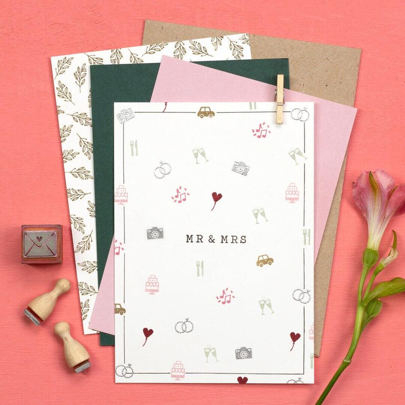 May include: A collection of wedding stationery, including a white card with "MR & MRS" text and wedding-themed icons, surrounded by envelopes and stamps. The color palette includes pink, green, and brown tones, with a pink background.