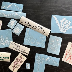 Peut inclure: Divers autocollants en vinyle blanc sur fond sombre. Les motifs incluent des cha&icirc;nes de montagnes, des motifs floraux, une main de squelette et une silhouette de Bigfoot. Certains autocollants portent les inscriptions "Best Wishes, Warmest Regards" et "Mountains Base."