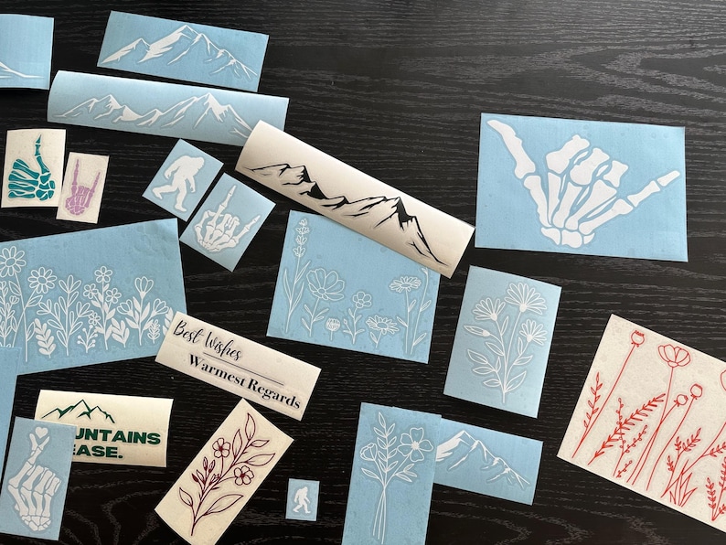 Peut inclure: Divers autocollants en vinyle blanc sur fond sombre. Les motifs incluent des cha&icirc;nes de montagnes, des motifs floraux, une main de squelette et une silhouette de Bigfoot. Certains autocollants portent les inscriptions "Best Wishes, Warmest Regards" et "Mountains Base."