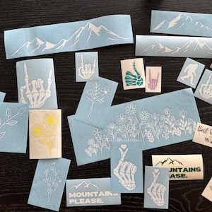 Peut inclure: Divers autocollants en vinyle bleu clair et cr&egrave;me avec divers motifs. Les autocollants pr&eacute;sentent des cha&icirc;nes de montagnes, des motifs floraux, des mains de squelette et le texte &laquo;MOUNTAINS PLEASE&raquo;. Certains autocollants ont le texte &laquo;Best Wishes, Warmest Regards&raquo;.