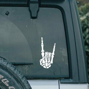 May include: A white skeleton hand making the rock and roll hand gesture. The hand is on the back window of a silver car.