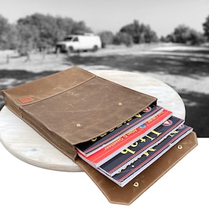 May include: A brown waxed canvas portfolio with a flap closure, holding several atlases. The portfolio has a small rectangular patch on the front and is secured with brass snaps. The atlases have red, black, and white covers.