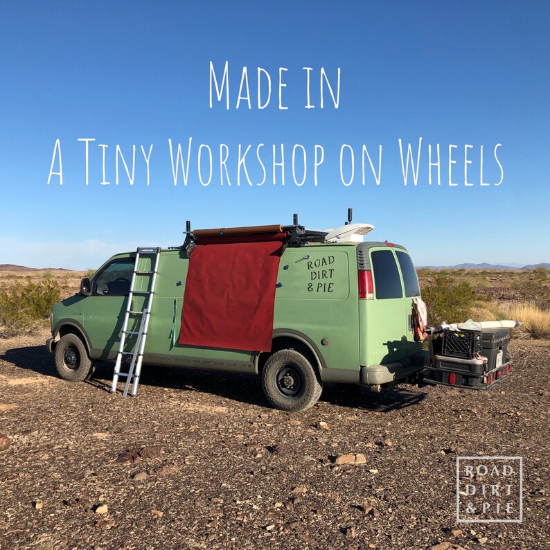 A green van with red waxed canvas draped down the side. The van has a ladder on the side and a sticker on the back that says 'Road Dirt & Pie'. The van is parked in the desert with a blue sky in the background. The text 'MADE IN A TINY WORKSHOP ON WHEELS' is superimposed on the image.