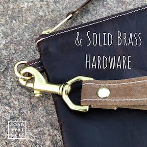 A close-up of a brown leather bag with a canvas strap and a brass clasp. The strap has a white stitch detail and a brass rivet. The text on the bag reads '& SOLID BRASS HARDWARE'.