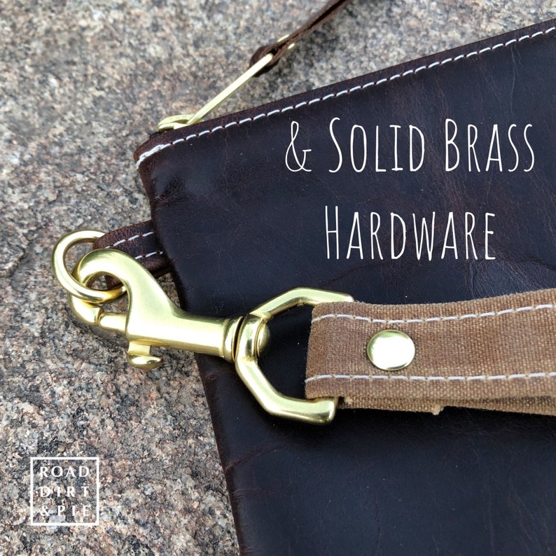 A close-up of a brown leather bag with a canvas strap and a brass clasp. The strap has a white stitch detail and a brass rivet. The text on the bag reads '& SOLID BRASS HARDWARE'.