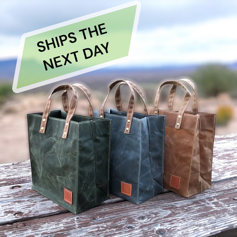 Three small tote bags on a weathered picnic table. Made with waxed canvas, they stand up on their own, and are naturally weather resistant. Color options are displayed as green, gray, and caramel brown. Handcrafted. Made in the USA.