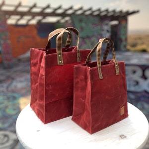 May include: Two red canvas tote bags with brown leather handles and brass accents. The larger bag is in the background and the smaller bag is in the foreground.