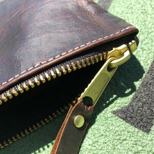 K&ouml;nnte beinhalten: Nahaufnahme einer braunen Leder-Rei&szlig;verschlusstasche mit einem goldenen Rei&szlig;verschluss und einem Messing-YKK-Rei&szlig;verschluss-Zipper. Die Tasche ist aus verwittertem Leder gefertigt und hat ein wei&szlig;es N&auml;hdetail.