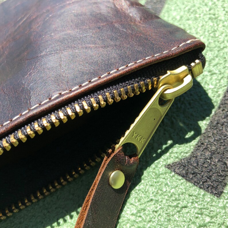 K&ouml;nnte beinhalten: Nahaufnahme einer braunen Leder-Rei&szlig;verschlusstasche mit einem goldenen Rei&szlig;verschluss und einem Messing-YKK-Rei&szlig;verschluss-Zipper. Die Tasche ist aus verwittertem Leder gefertigt und hat ein wei&szlig;es N&auml;hdetail.