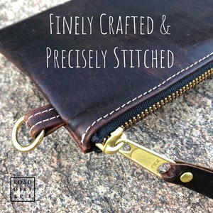 Close-up of a brown leather zipper pouch with white stitching. The zipper is brass with a YKK logo. The pouch has a small, brass ring on the side. The text 'FINELY CRAFTED & PRECISELY STITCHED' is visible above the pouch.