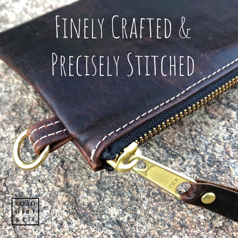 Close-up of a brown leather zipper pouch with white stitching. The zipper is brass with a YKK logo. The pouch has a small, brass ring on the side. The text 'FINELY CRAFTED & PRECISELY STITCHED' is visible above the pouch.