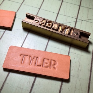 May include: A brass stamp with the letters "TYLER" is used to create a leather label with the same text.