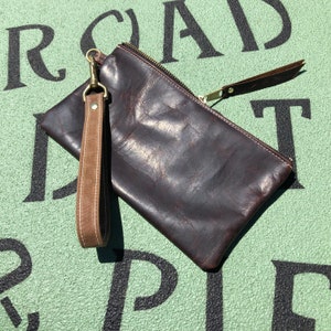 K&ouml;nnte beinhalten: Eine braune Leder-Clutch mit Rei&szlig;verschluss und einem braunen Lederriemen mit einem Messingverschluss. Die Clutch liegt auf einer gr&uuml;nen Oberfl&auml;che mit schwarzen Buchstaben.