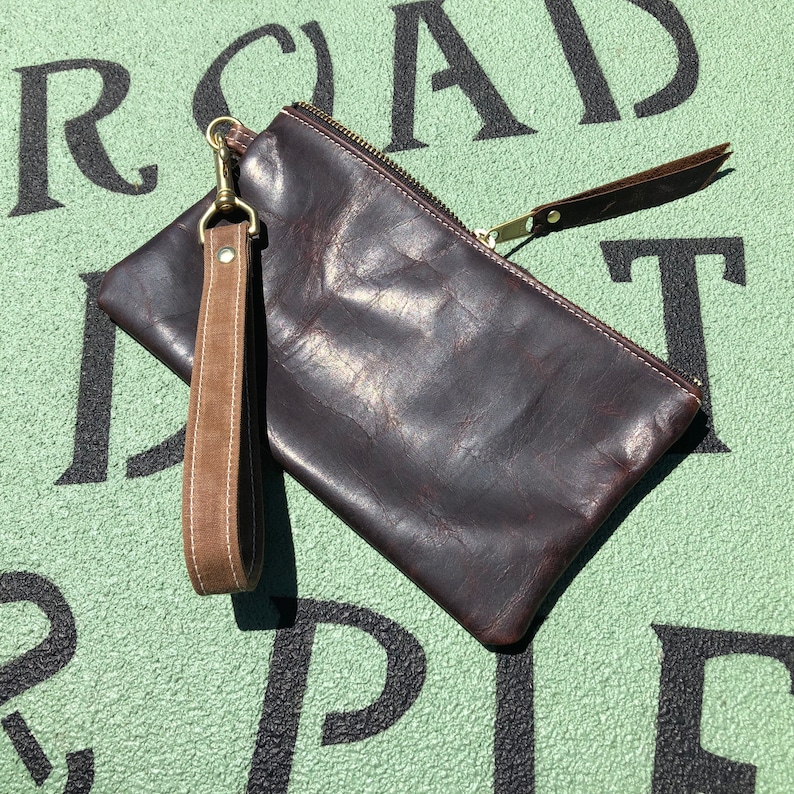 K&ouml;nnte beinhalten: Eine braune Leder-Clutch mit Rei&szlig;verschluss und einem braunen Lederriemen mit einem Messingverschluss. Die Clutch liegt auf einer gr&uuml;nen Oberfl&auml;che mit schwarzen Buchstaben.