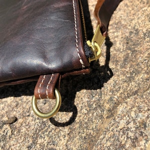 K&ouml;nnte beinhalten: Nahaufnahme einer braunen Ledertasche mit wei&szlig;en N&auml;hten. Die Tasche hat einen Messing-Rei&szlig;verschluss und einen Messingring am Riemen.
