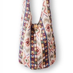 May include: A cream-colored shoulder bag with a colorful geometric pattern. The bag features a repeating design of diamonds, triangles, and floral motifs in shades of red, blue, yellow, and green. A wooden button secures the bag's opening.