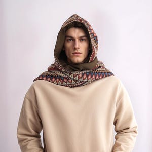 May include: A person wearing a beige hoodie with a patterned hooded scarf. The scarf has a geometric design in shades of red, brown, blue, and white.