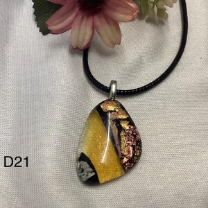 Peut inclure: Un pendentif en verre dichroïque sur un collier en cordon noir. Le pendentif a une forme unique, avec des teintes jaunes, noires et roses. Le collier est présenté sur une surface blanche, avec une fleur rose en arrière-plan. Le texte "D21" est visible.
