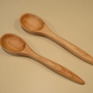 May include: Two light brown wooden spoons with smooth, rounded bowls and tapered handles. The spoons feature a visible natural wood grain pattern throughout their surfaces, showcasing a consistent light tan color and a polished finish.