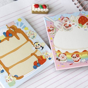 May include: Two sets of pastel-coloured memo pads featuring a cake design with animal characters. One pad has a light blue background with a multi-layered cake decorated with whipped cream, strawberries, and blueberries. The other pad has a pink background with a single-layer cake decorated with whipped cream, strawberries, and sprinkles. Both pads have a white writing area.