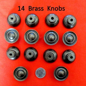 May include: Fourteen brass knobs with a dark finish are arranged on a red background. Each knob has a rounded, domed face and a central screw hole. The text "14 Brass Knobs" is at the top. A quarter is included for size comparison.