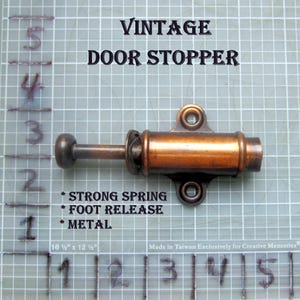 May include: A vintage bronze door stopper with the text "VINTAGE DOOR STOPPER". The door stopper has a strong spring and foot release. The door stopper is made of metal. The measurements are 10 1/2" x 12 1/2".