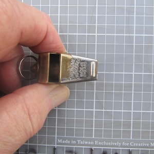 May include: A brass and silver-toned whistle with a ring for attachment. The text "THE ACME THUNDERER MADE IN ENGLAND" is printed on the side. The whistle is held in a hand.