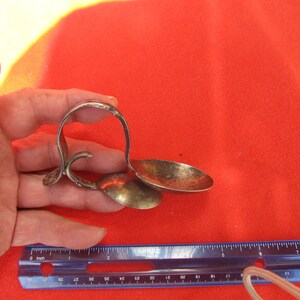 May include: A silver-toned spoon rest with two spoon holders and a curved handle. The spoon rest is held in a hand against a red background. A ruler is in the foreground.