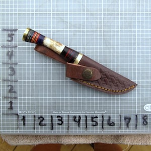 May include: A handcrafted knife with a decorative handle featuring red, blue, and black accents. The knife is paired with a brown leather sheath with a woven pattern and a brass button closure. The blade is not visible.