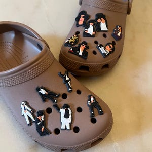 May include: Two brown clogs decorated with various black and white character charms. The charms depict a famous musician in different poses and outfits. The clogs are made of a rubbery material.