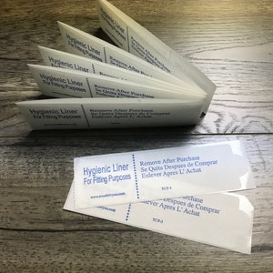 May include: Several white "Hygienic Liner For Fitting Purposes" liners are fanned out on a wooden surface. Each liner has text in blue, including "Remove After Purchase". The liners are for fitting purposes.
