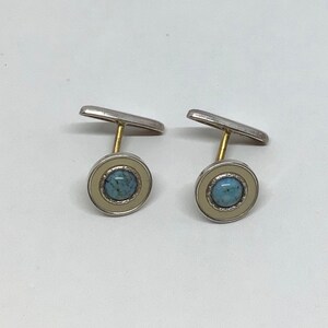 Puede incluir: Un par de gemelos plateados con una piedra central azul claro y un borde color crema. Los gemelos tienen un diseño clásico y están colocados sobre un fondo blanco.