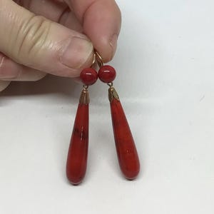 Könnte beinhalten: Ein Paar rote Ohrhänger mit goldfarbenen Akzenten. Jeder Ohrring hat eine kleine, runde Perle oben und einen langen, tropfenförmigen Anhänger. Die Ohrringe werden von einer Hand vor weißem Hintergrund gehalten.