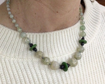 May include: A beaded necklace featuring a mix of light green and clear beads, interspersed with black and green accents. The necklace is worn over a white sweater.