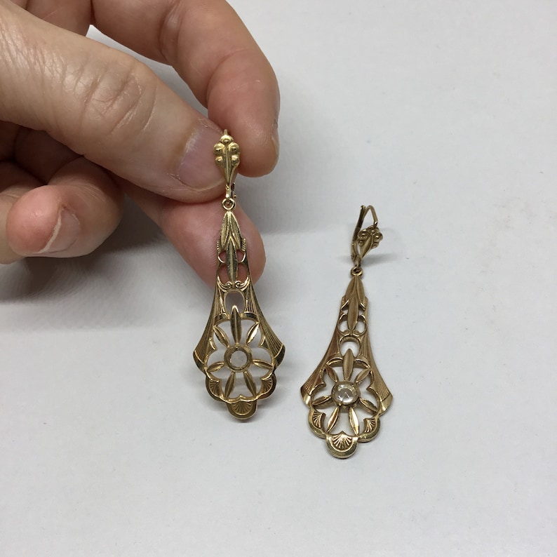 May include: A pair of gold dangle earrings with a delicate floral design and a clear stone in the center of each flower.