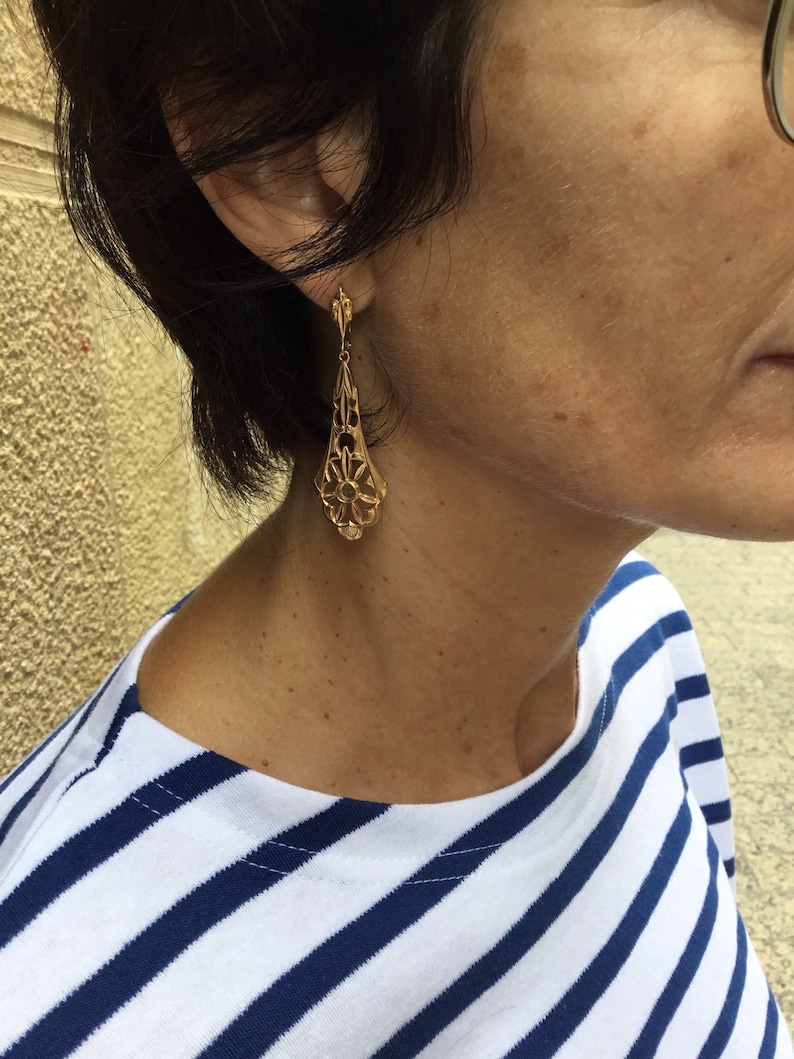 May include: Gold dangle earrings with a delicate, intricate design. The earrings are teardrop shaped and feature a floral pattern.