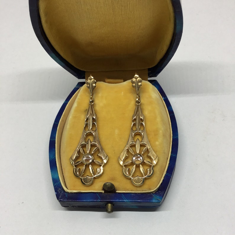 May include: A pair of gold dangle earrings with a clear stone in the center of each earring. The earrings are in a blue and gold jewelry box.