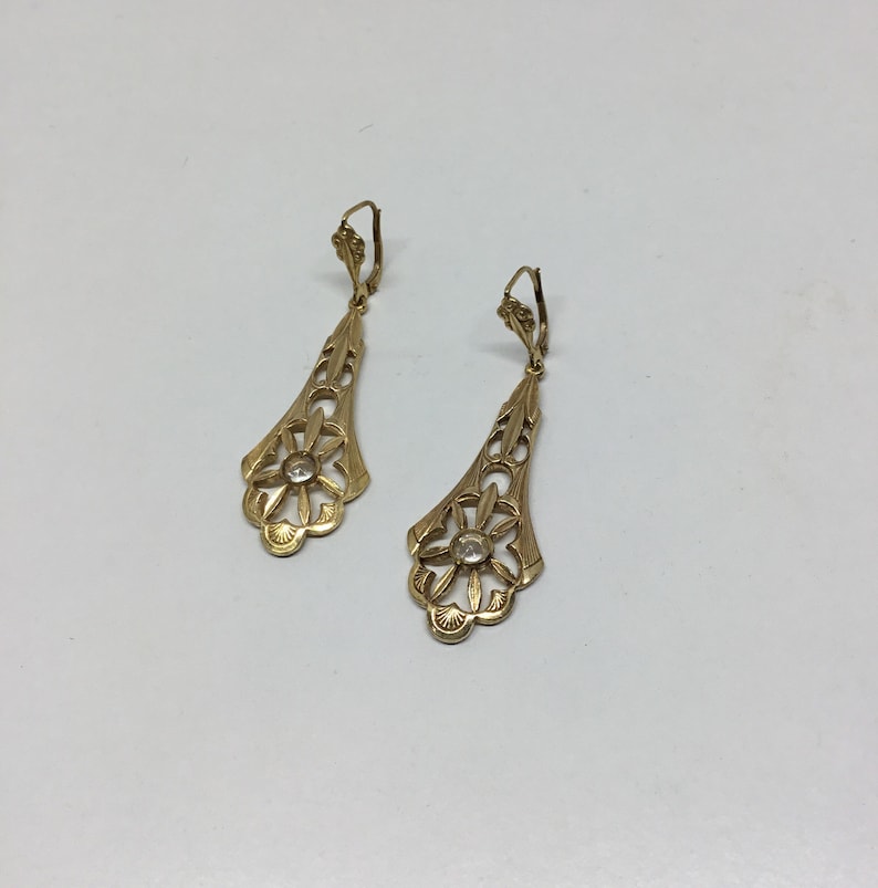May include: A pair of gold dangle earrings with a floral design and a clear stone in the center of each flower.