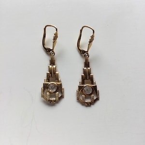 Könnte beinhalten: Ein Paar Ohrhänger im Art-Deco-Stil. Die goldfarbenen Ohrringe haben ein geometrisches Design und einen klaren, runden Edelstein in der Mitte. Die Klappbügelverschlüsse sind sichtbar. Präsentation vor weißem Hintergrund.