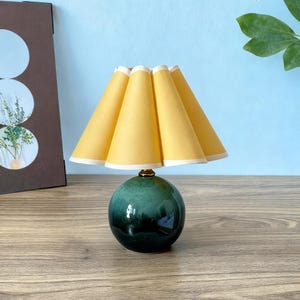 May include: A small, decorative table lamp with a yellow lampshade and a dark green, spherical base. The lampshade has a pleated design with white trim. The base has a glossy finish and a brass-colored accent at the top.